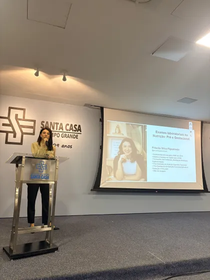 Priscila Figueiredo em pé, com microfone, apresentando diante de uma tela de conteúdo sobre exames laboratoriais na nutrição pré e gestacional